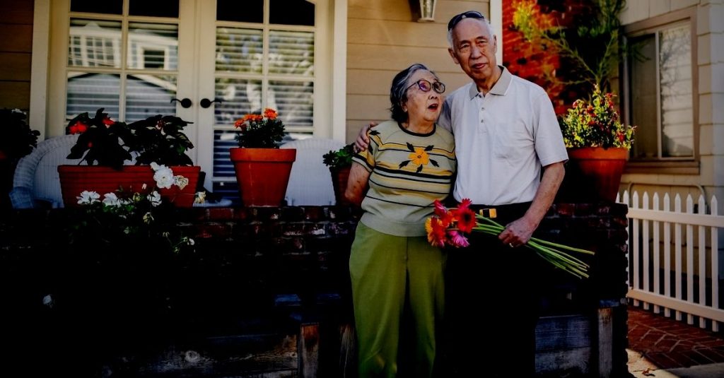 Asian elderly couple standing outside home. Man with flowers